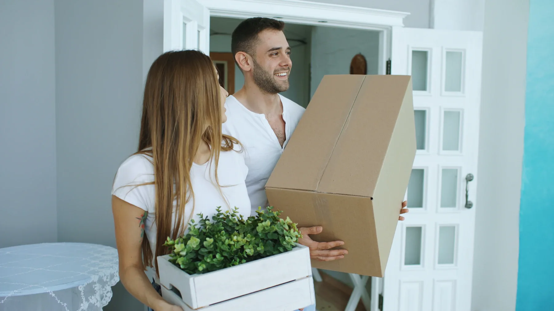 Young couple moving into new home carrying cardboard box and potted plant