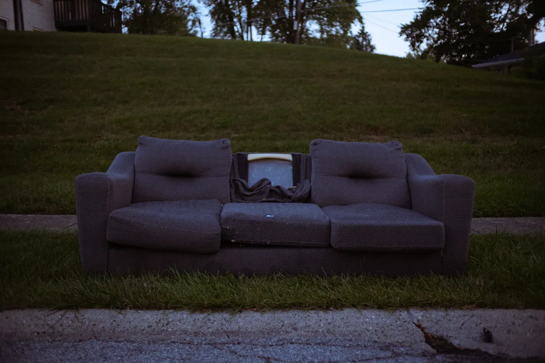 Abandoned dark purple couch sitting on grass beside a sidewalk with houses and trees in background